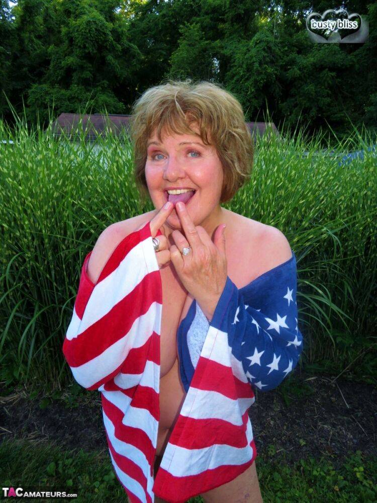 Older plumper Busty Bliss holds an American flag while totally naked in a yard - #14