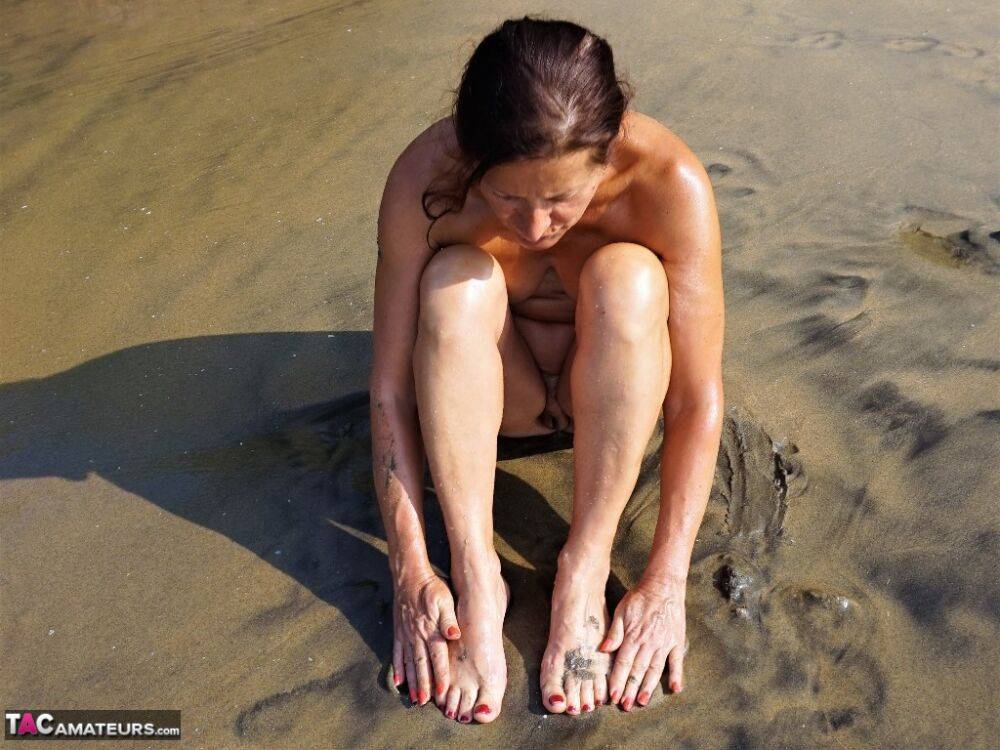 Naked older woman Diana Ananta covers her feet in beach sand at low tide - #5