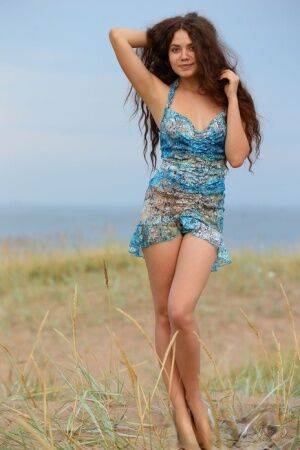 Long haired teen Norma A removes a dress to pose nude in a field near water on nudesceleb.com