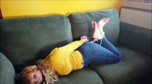 Barefoot white girl is hogtied on a sofa while ball gagged in her clothing on nudesceleb.com